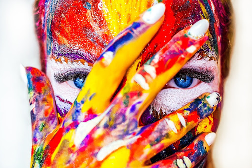 This is a color photography of a person's face. The person has red, yellow, white, blue, and purple paint splattered across their face, and the person is covering much of their face, with the exception of their eyes, with their right hand, which is also covered in paint.