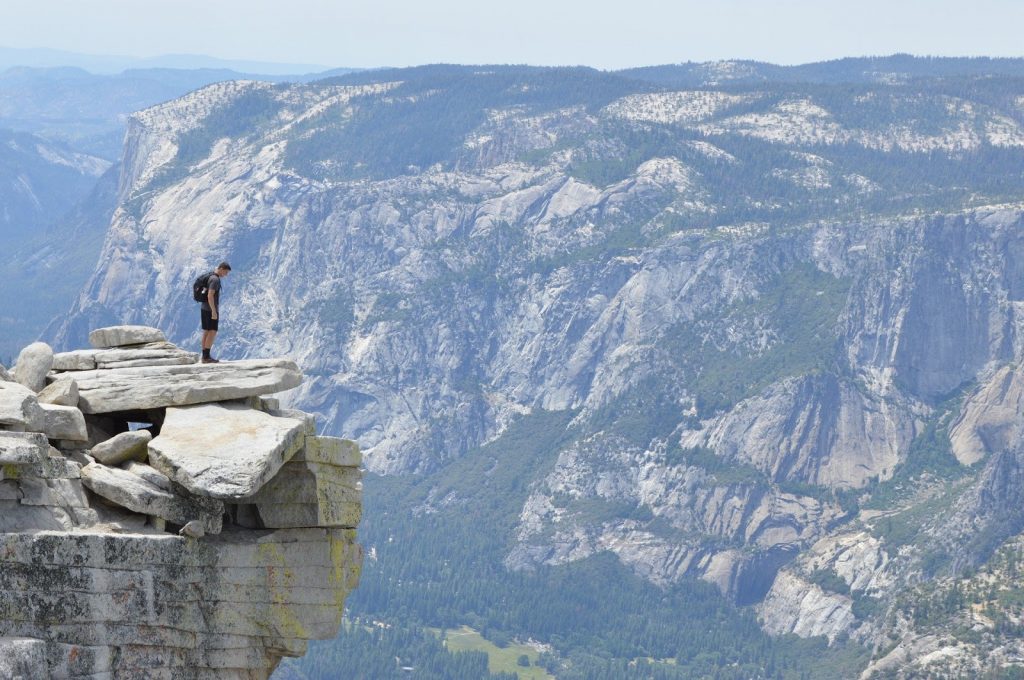 This is a color photograph of a man standing on top of a rock cliff overlooking a steep valley.