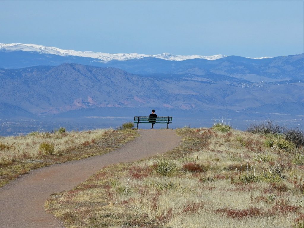 This is a color photograph of a person sitting alone on a bench outside. The bench is located off of a dirt path surrounded by a grassy field. Snow capped mountains are visible in the distance.