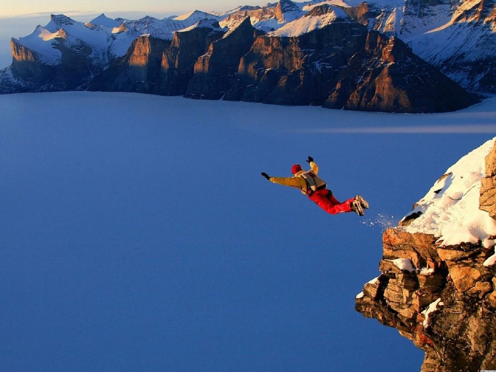 This is a color photograph of a person base jumping off of a cliff. The person is floating in mid-air above the snow-covered land. Snowy mountains are pictured in the distance.