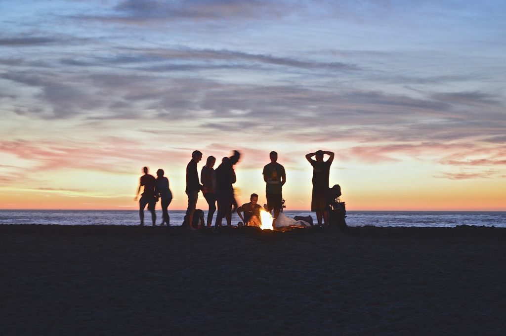 This color photograph shows eight people sitting and standing around a campfire by the ocean.