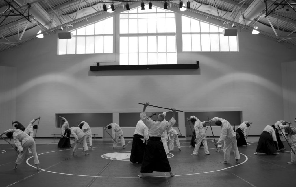 This is a black-and-white photograph of a group of adults stretching before an Aikido lesson.