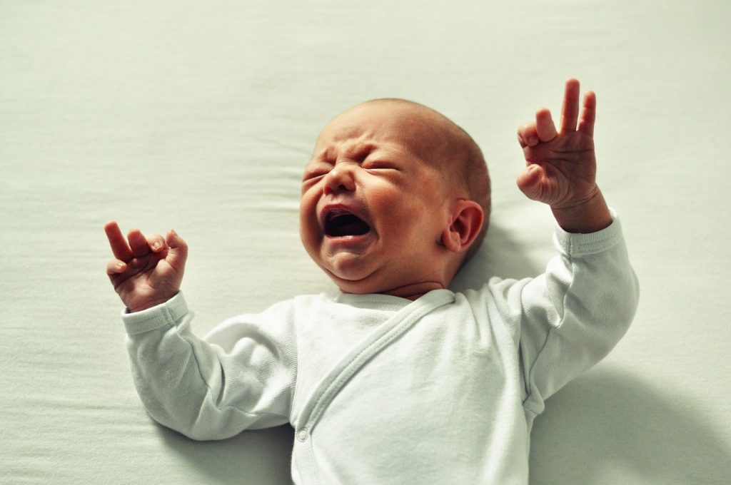This is a color photograph of a crying infant. The baby is lying on it's back and it is wearing a white, long-sleeved top.