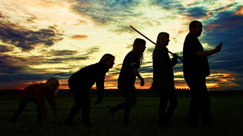 This is a color photograph of five people aligned in a row demonstrating the changes in human development through evolution. The person on the far left is walking on all fours; the next person in line is walking hunched over with his arms dangling in front; the next person in line is walking upright but with a curved back; the next person in line is walking upright while holding a stick across her shoulder; and the person on the far right is walking upright while looking down at the cell phone he is holding in his right hand.