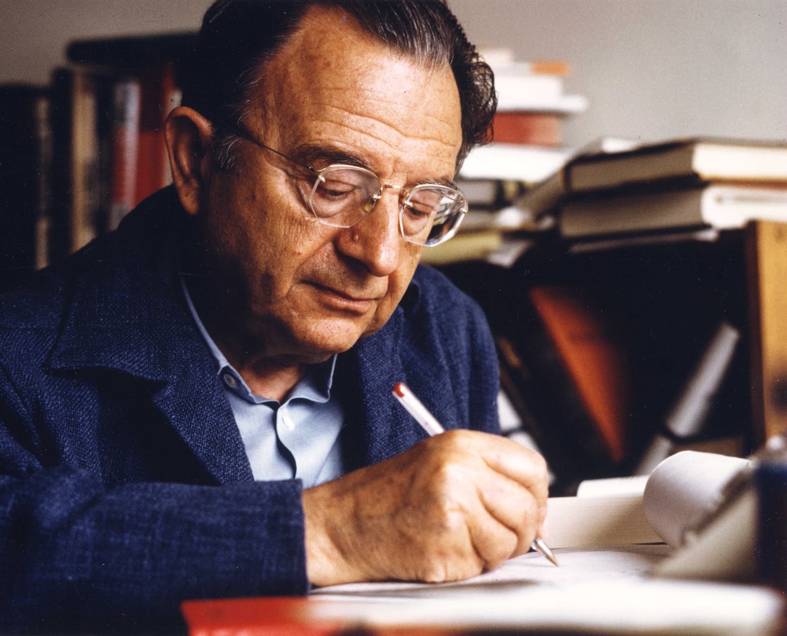 This is a color image of Erich Fromm taken in 1974. He is sitting at a desk writing on a piece of paper. A red pen is held in his right hand. He is wearing a blue shirt with a blue jacket on top. He is wearing eye glasses and he is looking down at the paper as he writes. There are books piled in the background.