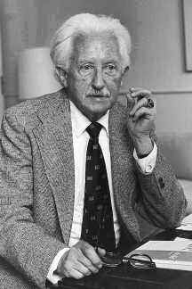 This is a black and white photograph taken of Erik Erikson. He is sitting in a chair as he stares past the camera. He is wearing a suit jacket, buttoned-up shirt, tie, and glasses. There are papers sitting on his desk.