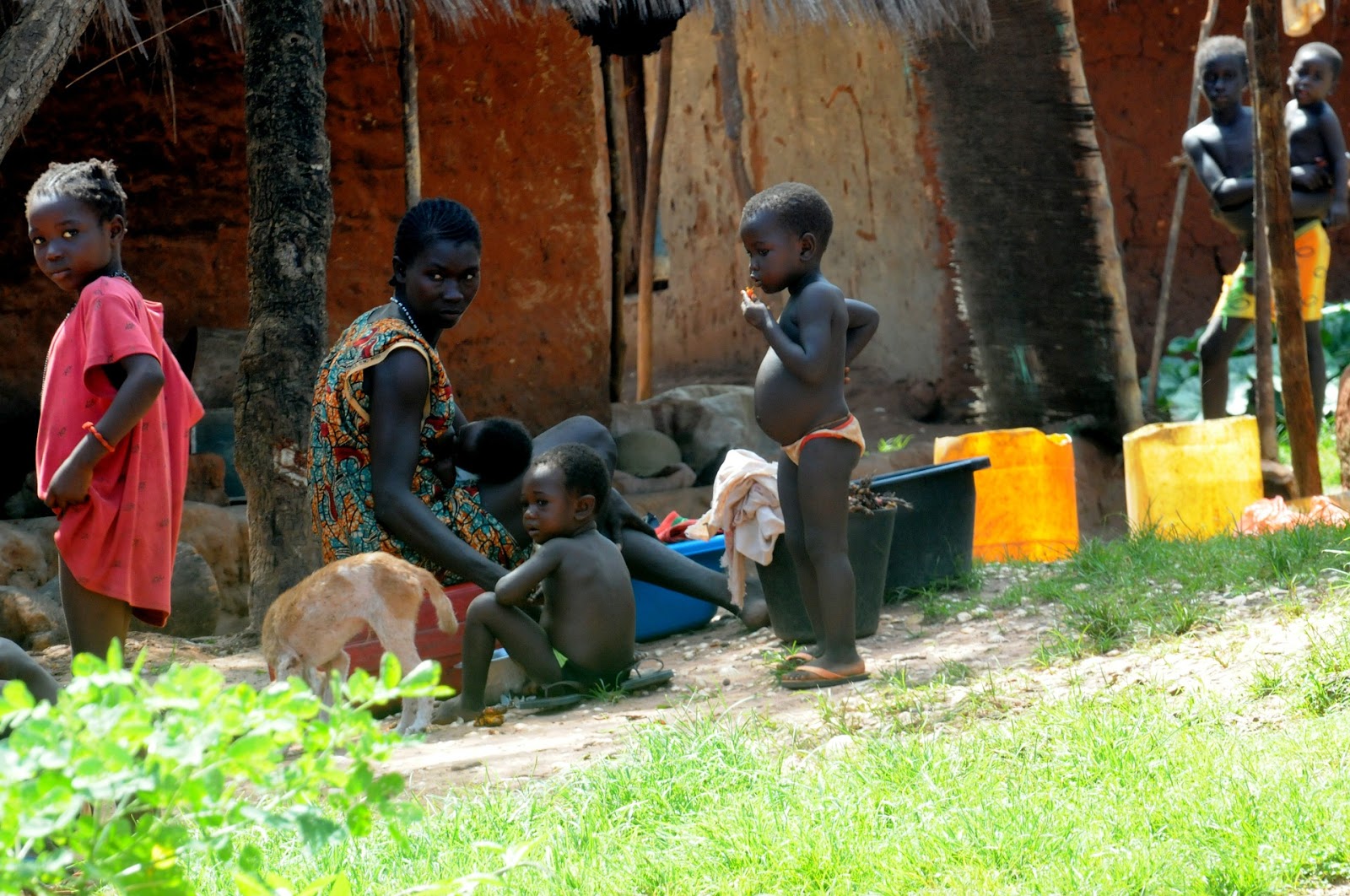 This is a color image of a Black mother and her four children. The family is outside of their home. The children are wearing few articles of clothing, and the mother is nursing her infant.