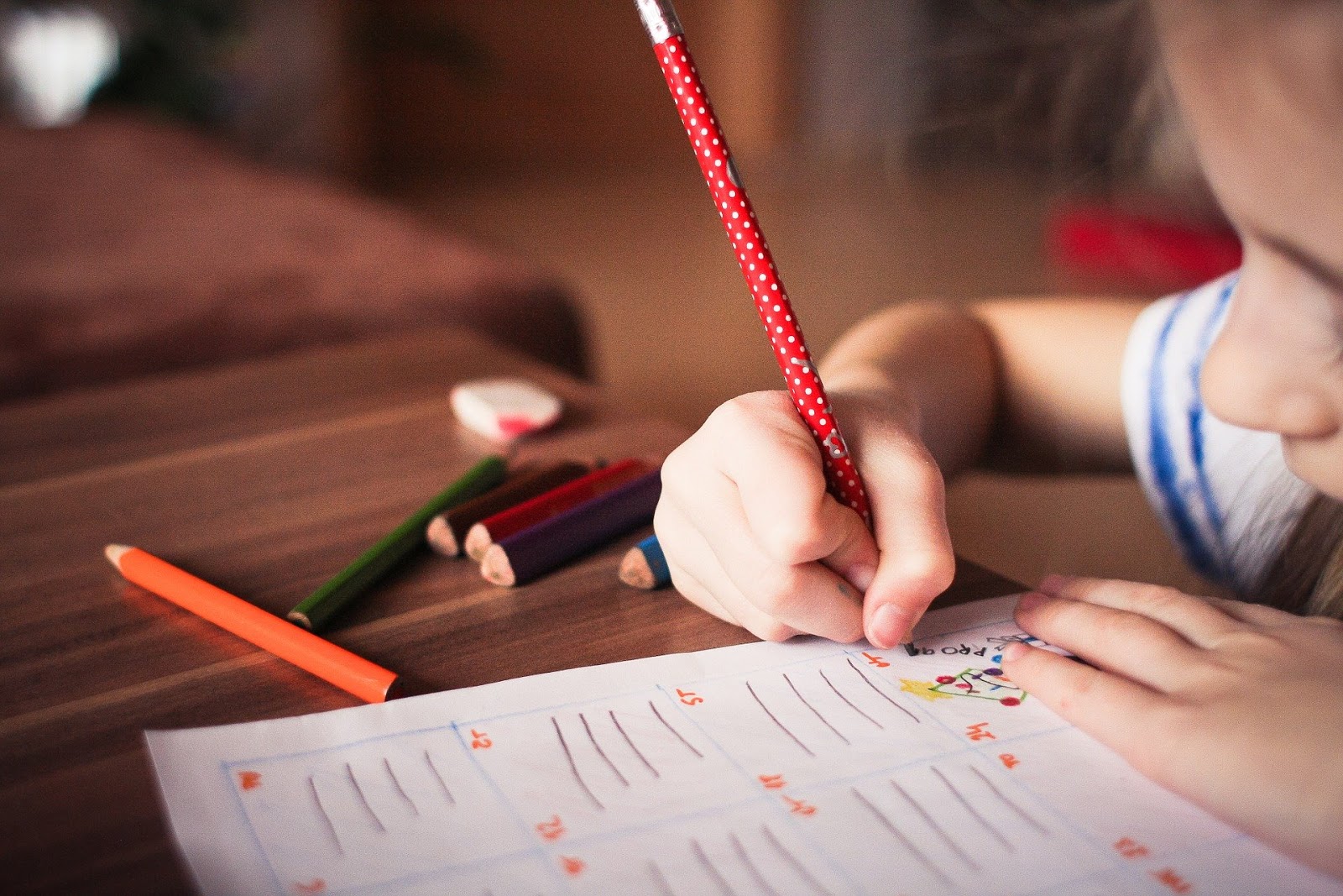 This is a color image of a child coloring on a piece of paper. There are several colored pencils on the table, and the child is holding a red pencil in their right hand.