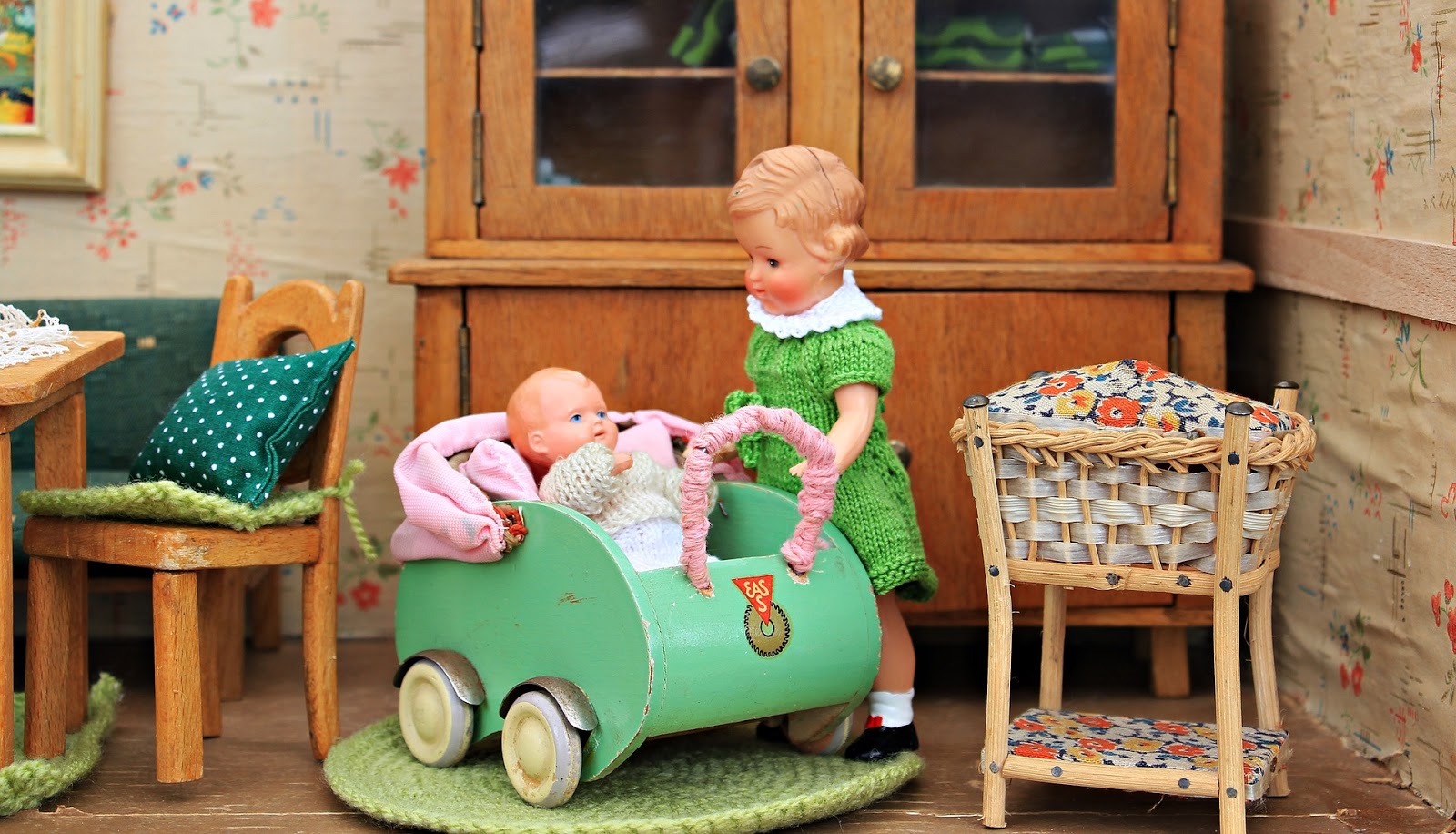 This is a color photo of two dolls in a dollhouse. One doll is standing beside a carriage, while the infant doll sits inside. There is dollhouse furniture pictured to the sides and rear of the dolls.