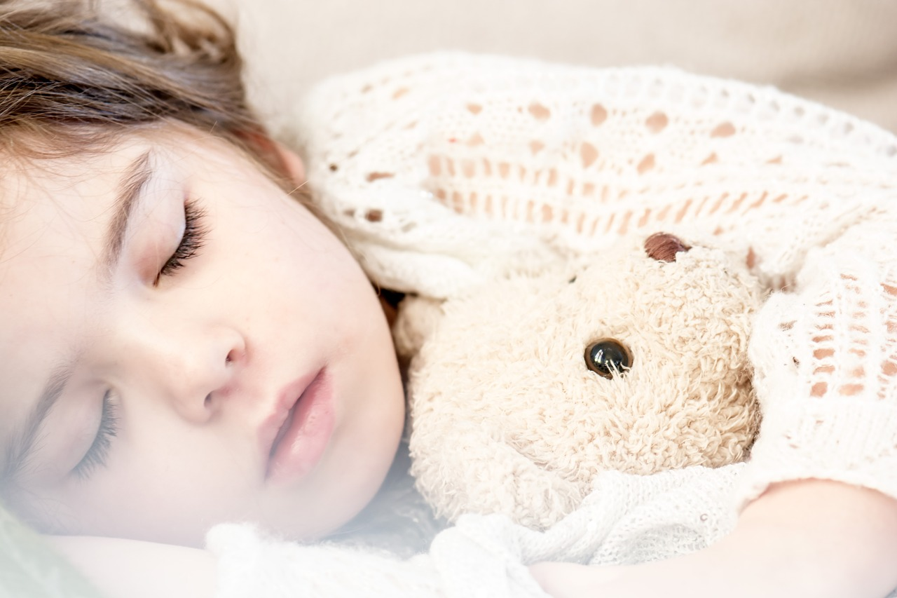 This is a color image of a young child taking a nap. The child has their arm wrapped around a white, stuffed teddy bear.