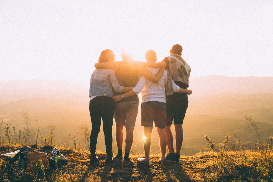 This is a color photograph of four adolescents standing in a row outside as they watch the sunset. They have their backs towards the camera, and they have their arms wrapped around one another's shoulders and waists.