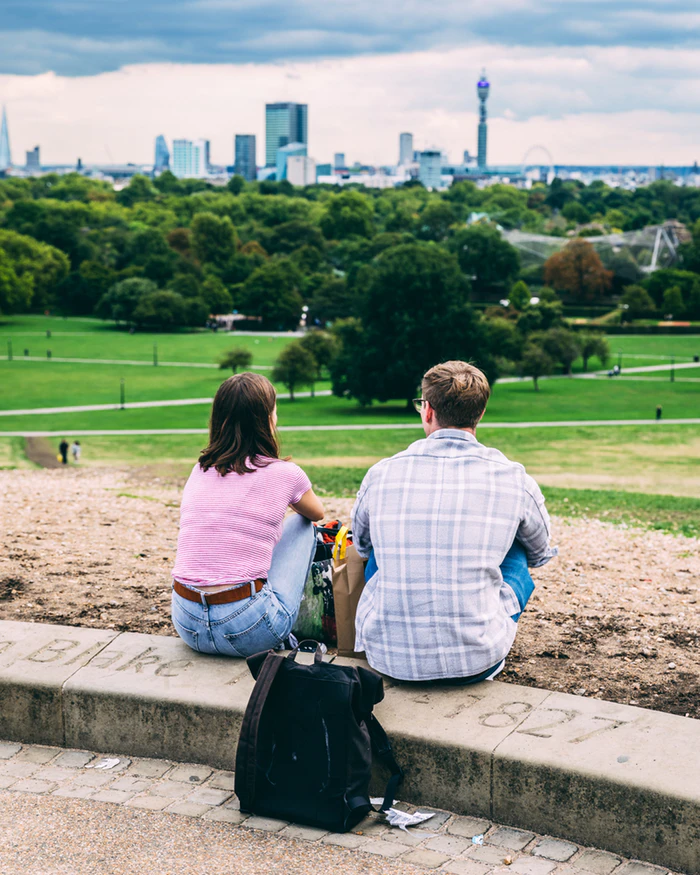 This is a color image of a man and woman sitting outside on a curb while looking out onto a field. A city skyline is visible in the distance. There is a backpack on the ground behind the couple.