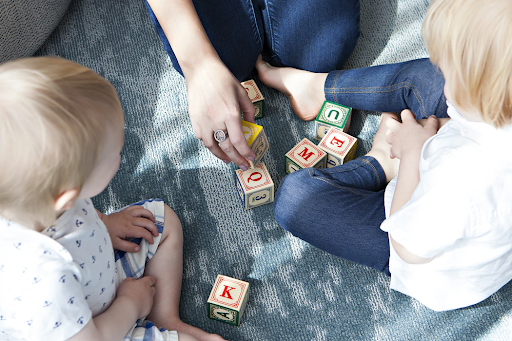 This is a color photograph of two toddlers sitting on the floor with an adult as they play with letter blocks.