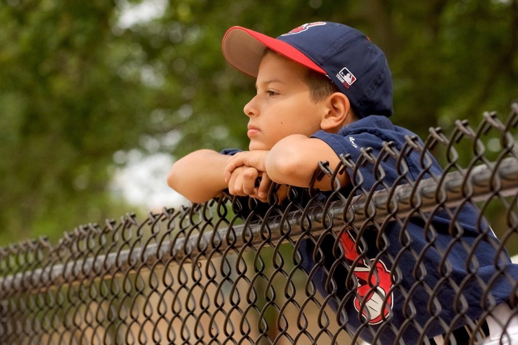 This is a color image of a young boy leaning on a chain-link fence. He is wearing a red and blue baseball hat and uniform top. The child is gazing out into the field.