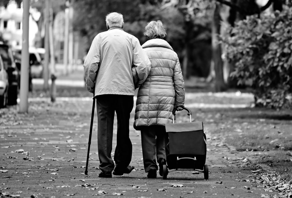 This is a black and white photograph of an elderly couple walking outside on a sidewalk. Their backs are facing their camera. The man is on the left and he is holding a cane in his left hand. The woman is on the right and she is pulling a suitcase with wheels using her right hand.