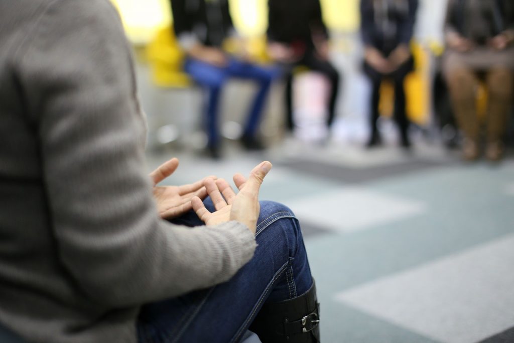 This is a color image of a person sitting in a chair. The image shows the person's legs and torso only. The person is resting his/her hands palm-up on their lap. There are people sitting in chairs in a circle visible in the background.