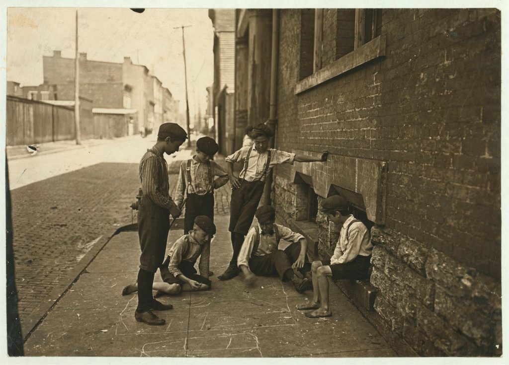 This is a black and white photo taken in 1908 of six boys hanging around outside on the sidewalk. There are two boys sitting on the ground drawing with chalk.