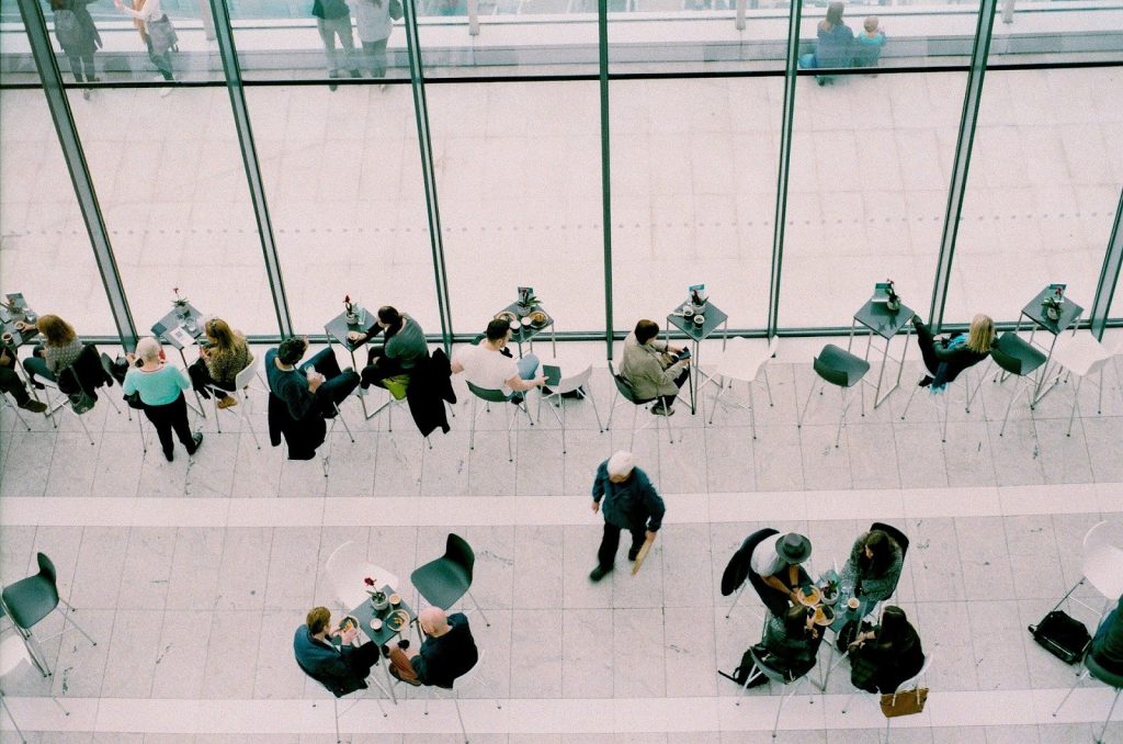 This is a color, aerial photograph of people sitting at tables in a glass atrium. The tables and chairs are spread out around the floor, and people are sitting in small groups. The people are dressed in business attire.