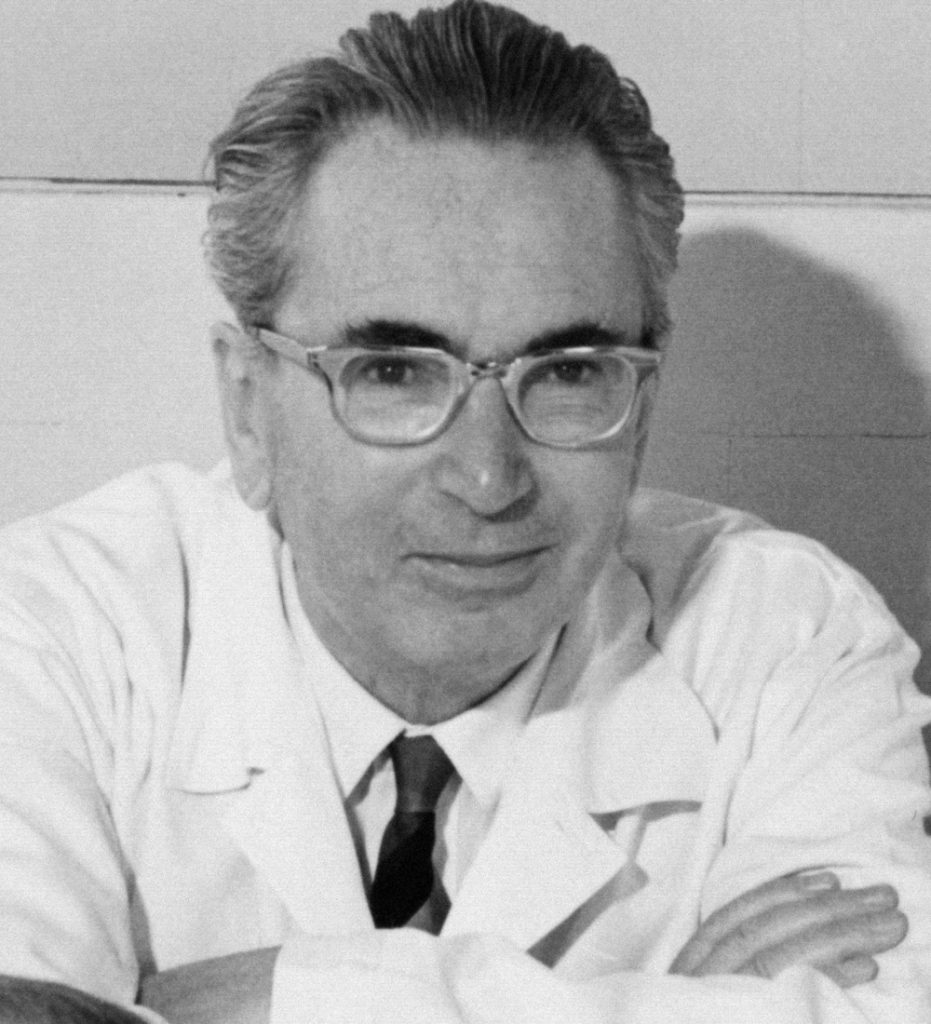 This is a black and white photograph taken of Viktor Frankl in 1965. He is wearing a white labratory coat, a collared shirt and a tie. He has his arms crossed out in front of him as he leans on a table to pose for the picture.