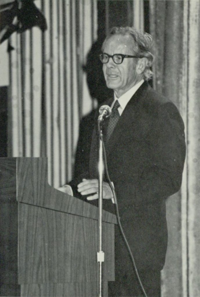 This is a black and white photograph taken of Rollo May giving a speech at a podium. He is dressed in a suit and tie and there is a microphone on a stand to his left.