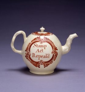 White teapot with red design and words, “Stamp Act Repealed.” The background of the image is purple.