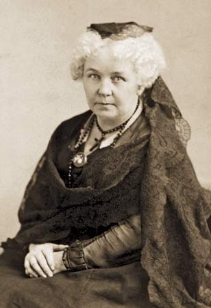 A photograph of Elizabeth Cady Stanton. She is wearing a dress, a hat, /and a cross around her neck.