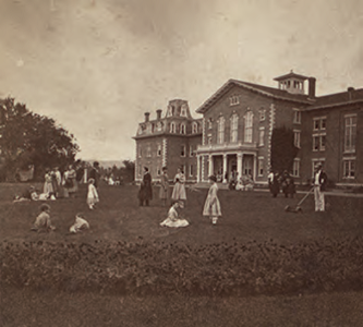 A black and white photograph of the main building of the Oneida community. A large house with pillars with men and women on the front lawn.