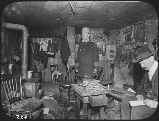 Two New York City Tenement House Department officials inspecting a cluttered basement living room in a tenement house, 1900.